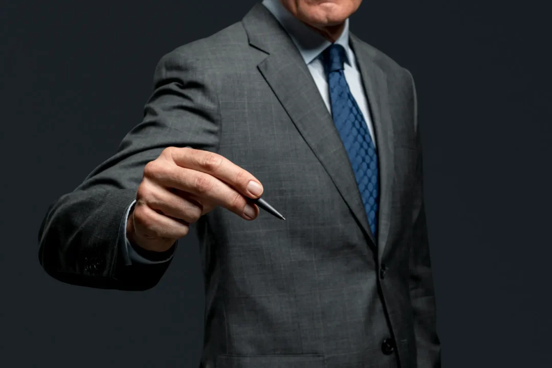 עורך דין נדלן מגיש עט לבוש בחליפה יוקרתית על רקע שחור כהה.